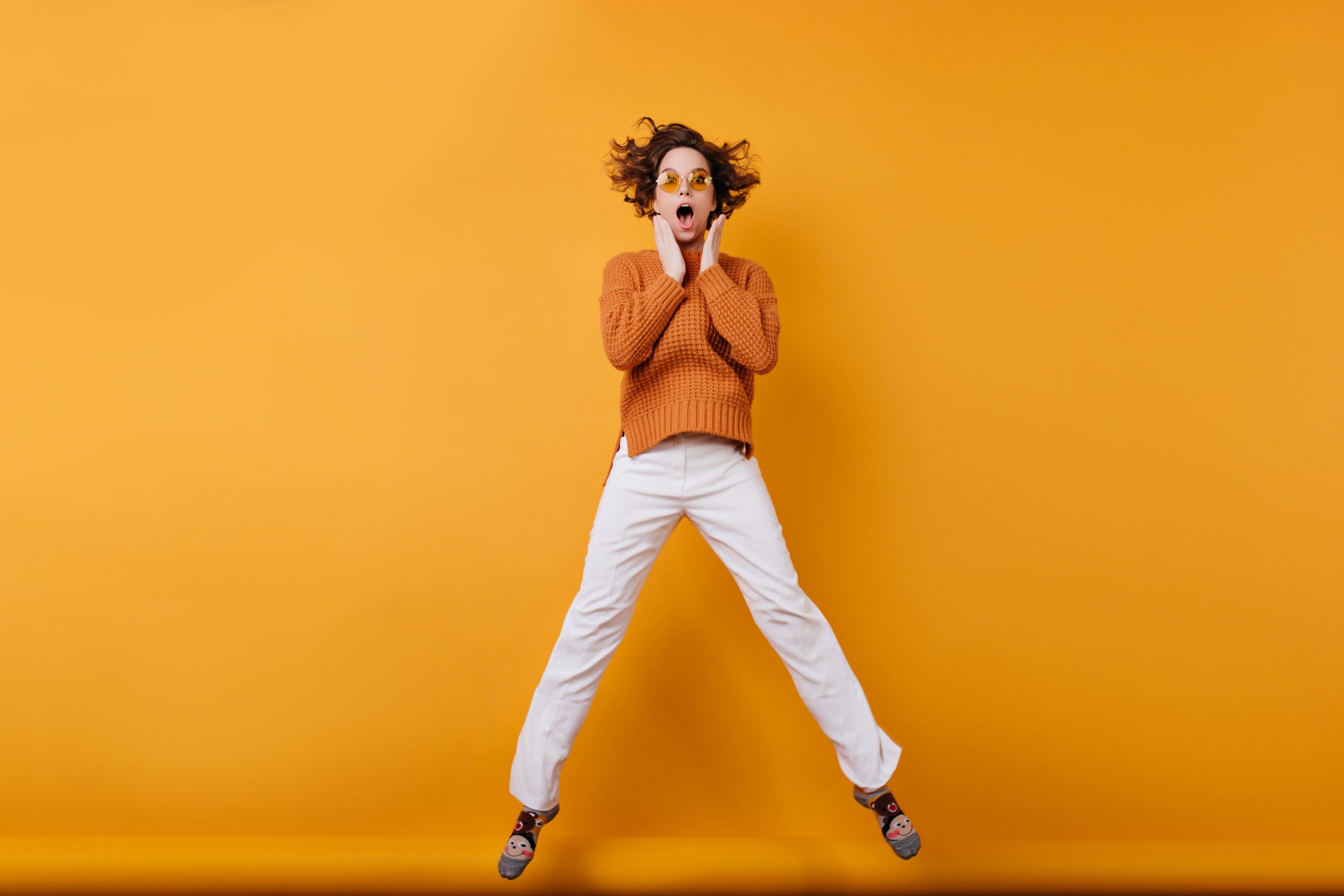 Full-length photo of amazed young woman in white pants dancing in studio. Portrait of wonderful girl with wavy hair jumping with funny face expression.